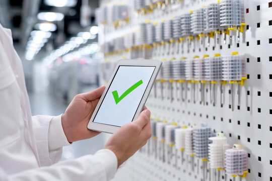 Technician in white lab coat holds tablet displaying green check mark while standing in front of organized wall of electrical components in a modern facility