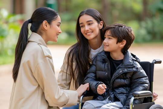 Two women interact with a young boy in a wheelchair, smiling and engaged in conversation in a park setting with greenery and pathways visible in the background