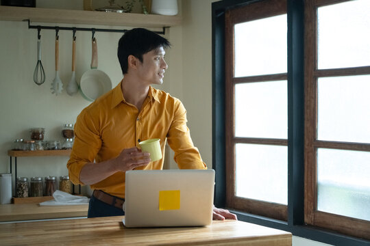 Young Asian Man Reading Morning News on Laptop with Coffee in Kitchen Home Office
