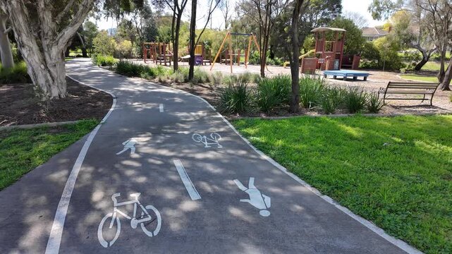 A paved shared user path winding through a leafy suburban park in Yarraville,&nbsp;with painted lane markings and icons for pedestrians, bicycles beside a children's playground. Melbourne, Australia.