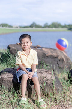 A cute young Asian boy sitting on a large rock by a calm lake with clasped hands, wearing a yellow polo shirt and cute denim shorts, smiling happily outdoors on a bright sunny day with a beach ball.