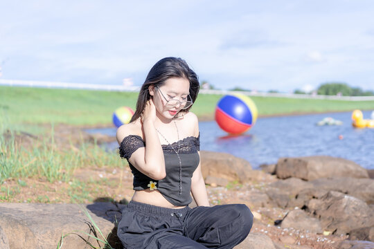 A relaxed young Asian woman sitting on a rock by a serene reservoir, touching her neck while wearing glasses and a fashionable black top with pants on a bright sunny day with a colorful floating ball.