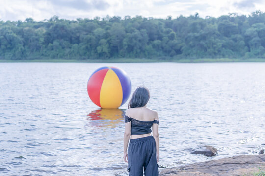 A young Asian woman standing with her back to the camera by a serene reservoir, looking out across the water wearing a stylish black outfit on a bright sunny summer day, with a colorful floating ball.