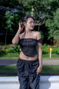 A thoughtful young Asian woman standing by a white metal guardrail in a scenic green park, touching her hair while wearing glasses and a fashionable black top and pants on a bright sunny summer day.