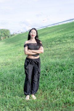 A smiling young Asian woman standing on a lush green hill by a serene reservoir with arms crossed, looking at the camera while wearing glasses and a stylish black outfit on a bright sunny summer day.