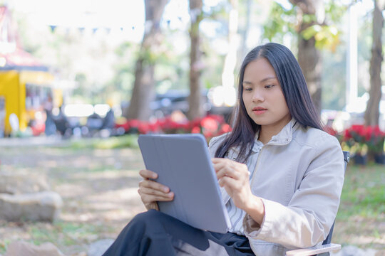 A focused young Asian woman sitting outdoors in a scenic park, holding a digital tablet and a stylus pen while checking a photo on the screen with a concentrated expression amidst lush green foliage.