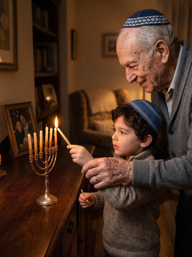 Elderly grandfather guides young grandson lighting traditional menorah candles during holiday at home. Hanukkah or Chanukah Jewish celebration concept with copy space.