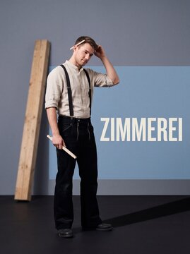 Zimmerer posiert mit Zollstock vor Holzbalken und Zimmerei Schriftzug