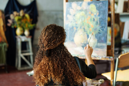 Teenage girl in college painting class, art studio, art workshop. She paints still life on canvas with oil paints, holds brush, palette.