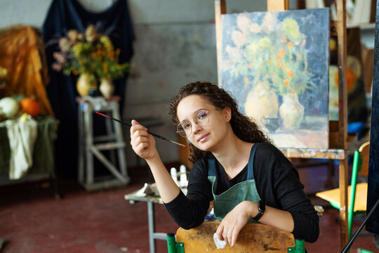 Portrait of teenage girl in painting class, art studio, art workshop. She paints still life on canvas with oil paints, holds brush, palette. Studying painting in college.