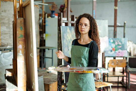 Portrait of teenage girl in painting class, art studio, art workshop. She paints still life on canvas with oil paints, holds brush, palette. Studying painting in college.