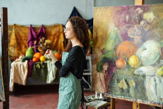 Teenage girl in college painting class, art studio, art workshop. She paints still life on canvas with oil paints, holds brush, palette.