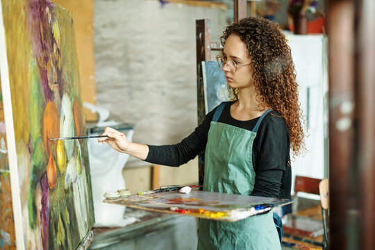 Teenage girl in painting class, art studio, art workshop. She paints still life on canvas with oil paints, holds brush, palette. Studying painting in college.