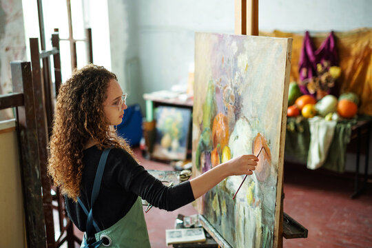 Teenage girl in painting class, art studio, art workshop. She paints still life on canvas with oil paints, holds brush, palette. Studying painting in college.