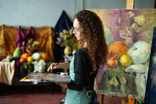 Teenage girl in painting class, art studio, art workshop. She paints still life on canvas with oil paints, holds brush, palette. Studying painting in college.