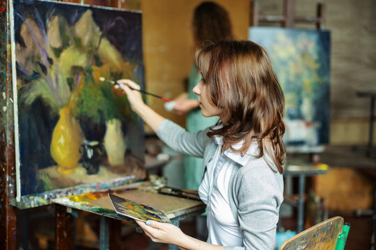 Teenage girl in painting class, art studio, art workshop. She paints still life on canvas with oil paints, holds brush, palette. Studying painting in college.