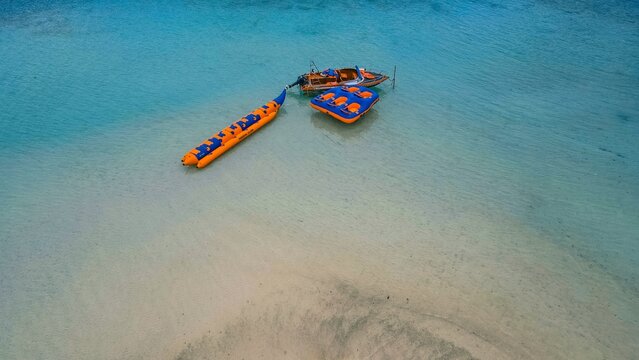 Aerial view of Pahawang Island featuring a banana boat and a water tube attached to a motorboat in the clear turquoise shallow waters of Kecamatan Punduh Pidada, Lampung, Indonesia.