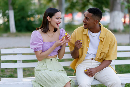 Young couple sitting on a park bench sharing conversation and snacks, dressed in casual summer attire, surrounded by greenery and soft natural light