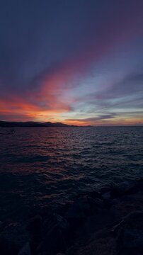 Vertical. Dark Ocean Waves Ripple Beneath a Colorful Sunset, Viewed From a Rocky Shore With Distant Hills, Evoking Tranquility And Natural Beauty Vertical video.