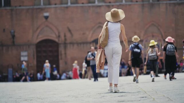 Stylish woman wearing light clothes and a wide-brimmed hat strolls through the sunlit Piazza del Campo in Siena, Tuscany, Italy