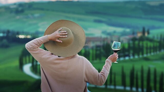 Picturesque scene of stylish woman standing in a Tuscany field, holding a glass of red wine with background of rolling hills and cypress lined road