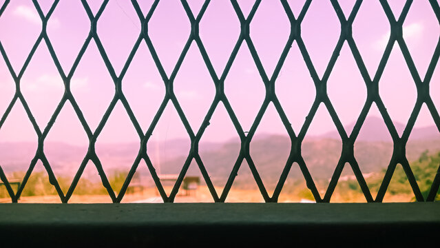View from inside a room through a dark green diamond-patterned metal trellis window, looking out towards a scenic landscape of hills and trees under a hazy pinkish sunset sky.
