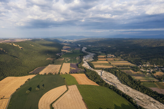 Aerial view of the Asse river winding through agricultural fields, lavender rows, and rolling hills under a cloudy sky in Bras-d'Asse, Provence-Alpes-Cote d'Azur, France.