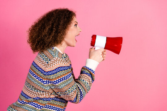 Woman shouting into a red megaphone on pink background wearing patterned sweater, energetic confident portrait communicating announcement and attention in studio