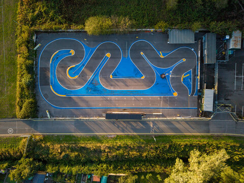 Aerial view of an RC racing track with asphalt surface, blue painted sections, and yellow curbs, surrounded by trees and greenery in Stafford, England, United Kingdom.