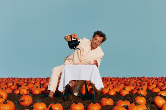 Man in a cream linen suit pouring tea at a formal table in a muddy pumpkin field, absurd seasonal scene with copy space for autumn hospitality