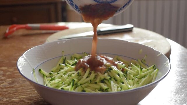 Close-up shows tomato dressing flowing from patterned bowl into large white ceramic bowl filled with shredded zucchini, creating glossy orange red ribbons across colorful vegetable mix in warm