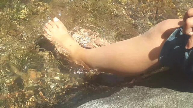 Someone was sitting on arock while soaking his feet in a clear and shallow stream of water