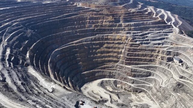 Aerial drone view of a vast open-pit mine with dramatic terraced levels, winding haul roads, exposed rock formations and heavy machinery across a rugged industrial landscape.