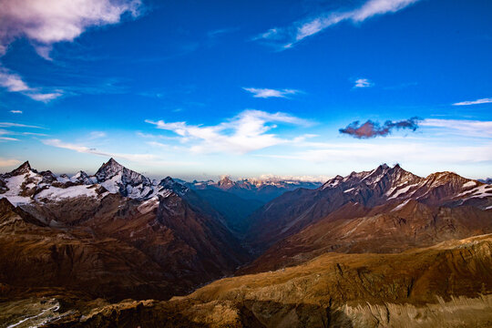 Aerial view of the majestic snow-capped peaks of the Alps surrounding the Zermatt valley under a bright blue sky with wispy clouds in Zermatt, Valais, Switzerland.