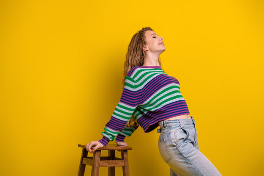 Young woman in colorful striped sweater leaning on a stool against a yellow background