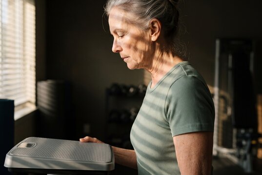 A mature woman holds a bathroom scale while standing in a home gym, contemplating her health and fitness progress