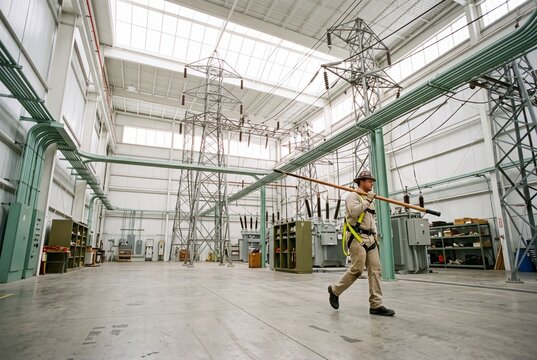 Electrical technician carrying a hot stick walks through an indoor high-voltage power substation facility