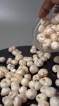 A hand pour crispy white lotus seeds Euryale ferox from a small glass bowl onto a dark plate surface for healthy snack food scene