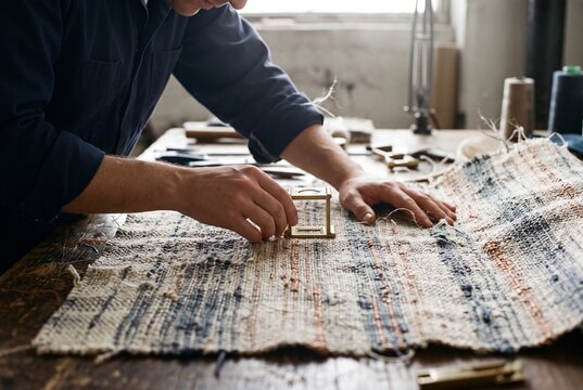 Textile artisan inspecting handwoven fabric with a brass magnifying loupe on a wooden workshop table
