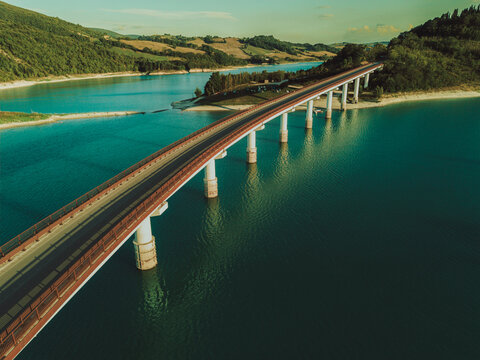 Aerial view of the bridge spanning Lago di Castreccioni surrounded by lush green hills and calm blue water in Cingoli, Marche, Italy.