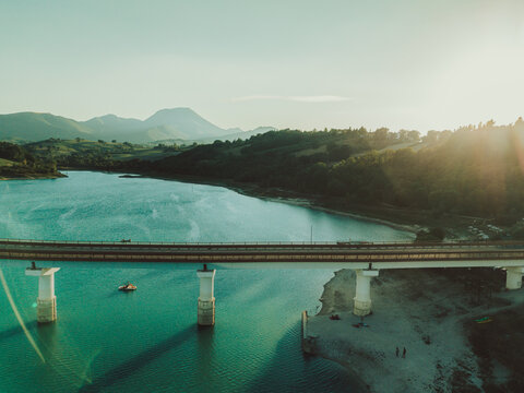 Aerial view of Lago Castreccioni with a bridge crossing the water, surrounded by rolling hills and lush greenery under the warm glow of the golden hour in Cingoli, Marche, Italy.