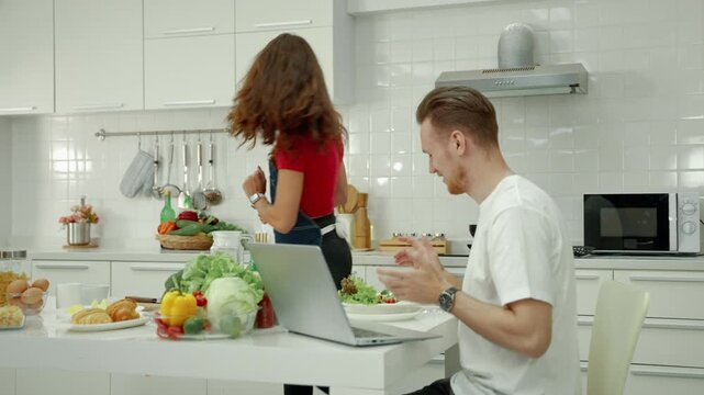 Woman serving a fresh healthy salad to her partner while he sits at the kitchen table with a laptop, surrounded by fresh vegetables and food.
