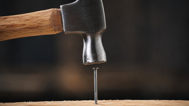A close-up of a metal hammer head hitting a steel nail driven into a wooden surface