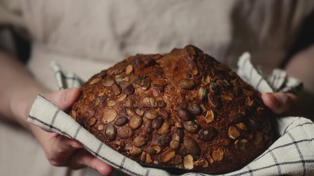 Close-up of baker in a rustic linen apron holding freshly baked artisan bread topped with pumpkin seeds on a checkered towel. Homemade loaf for bakery promos, organic product and healthy food concept.