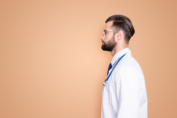 Profile portrait with copy space, empty place for advertisement of attractive stylish doc with stubble in white lab coat and stethoscope on his neck, isolated on grey background © deagreez
