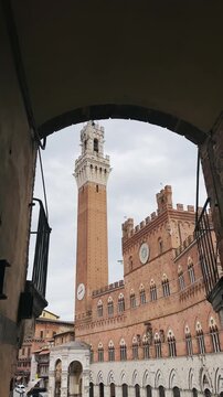 Historic Torre del Mangia and Palazzo Pubblico in Siena, Tuscany, captured on a cloudy day framed with narrow alley archway, vertical video