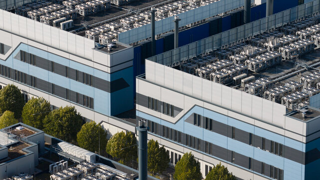 Aerial view of a modern industrial data center facility with rows of cooling units on the roof, blue and grey metal facade, and surrounding green trees in Frankfurt am Main, Hessen, Germany.