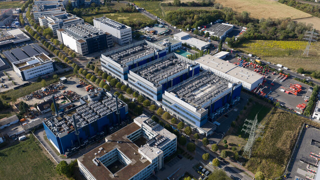 Aerial view of a large modern data center complex with extensive rooftop cooling infrastructure, surrounded by industrial buildings and green spaces in Frankfurt am Main, Hessen, Germany.
