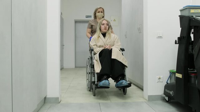 Doctor escorts paralyzed woman toward ultrasound examination room. Nurse guides wheelchair toward procedure area in modern hospital. Slow motion