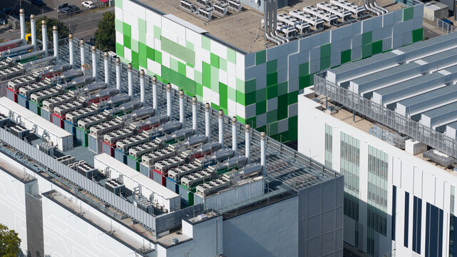 Aerial view of a modern data center facility featuring a green and white pixelated facade and rows of industrial cooling units on the roof in Frankfurt am Main, Hessen, Germany.